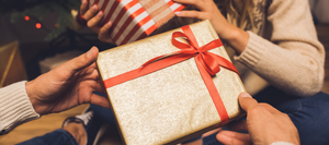 Close-up of two hands exchanging a wrapped gold gift box tied with a red ribbon. A blurred background shows a cozy holiday setting with a Christmas tree and another gift.